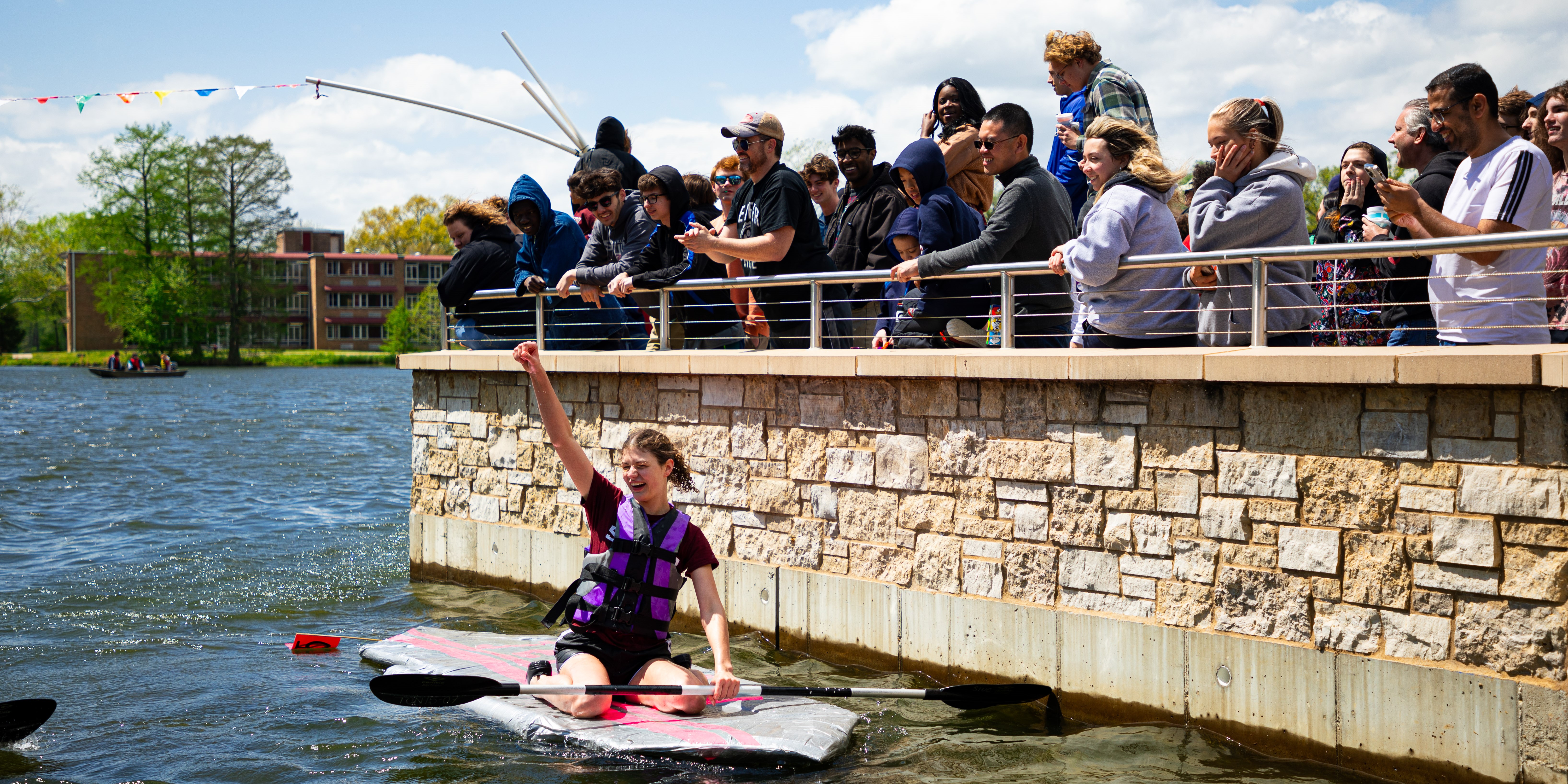 Crowd cheering for a victorious student on a paddleboard on Campus Lake.